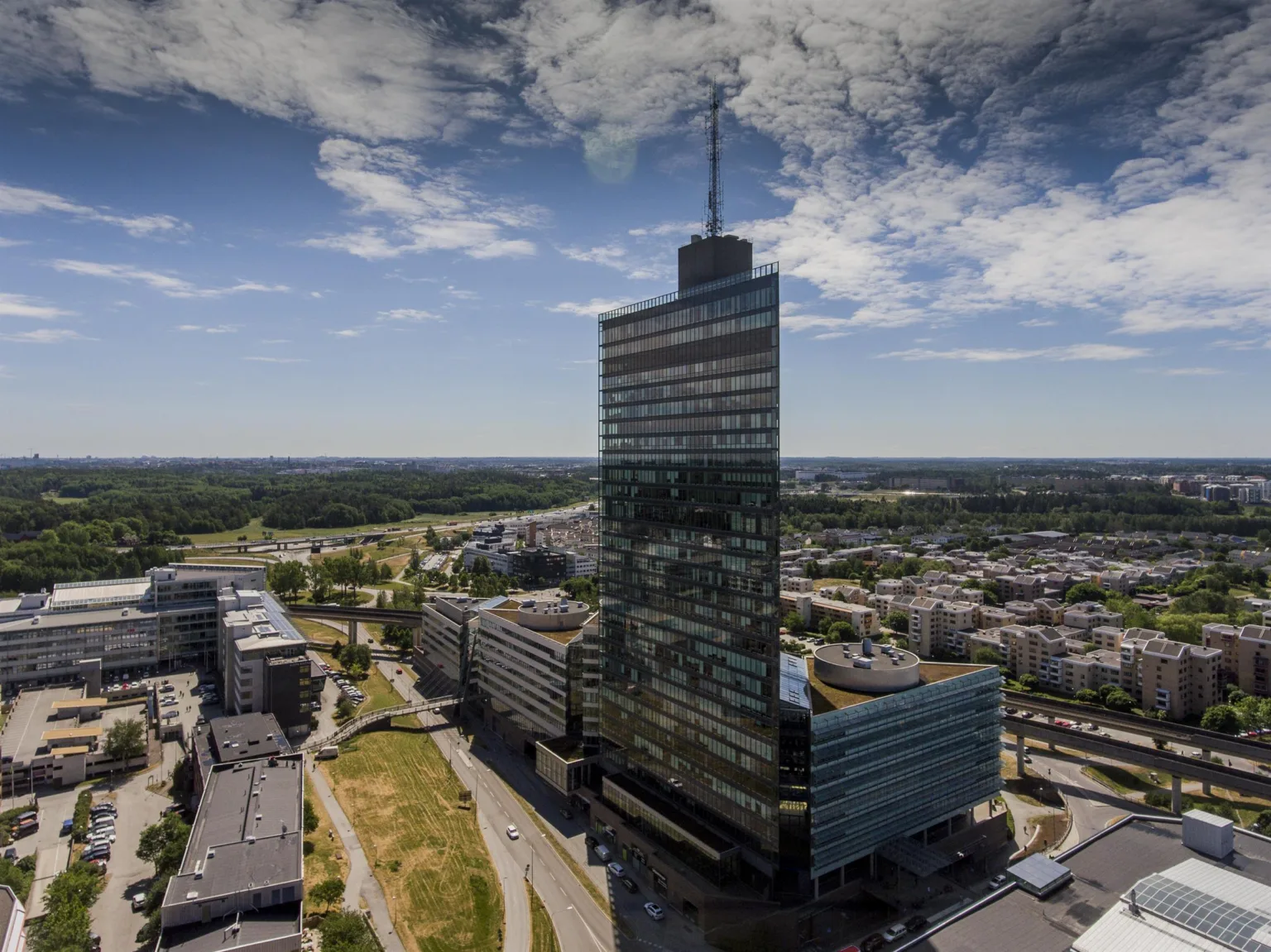 En hög skyskrapa med glasfasad omgiven av stadslandskap och grönområden under en blå himmel med moln.