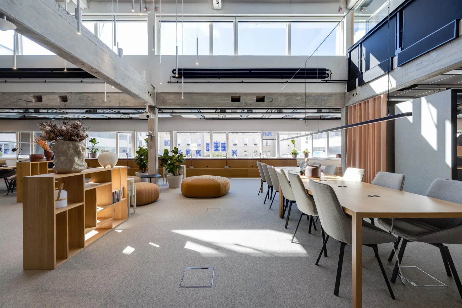 1. En modern arbetsplats med stora fönster, bord, stolar och sittpuffar samt växter placerade runt rummet.