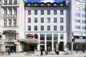 En vit byggnad med stora fönster, en entré med skyltar, och människor som står eller går utanför.