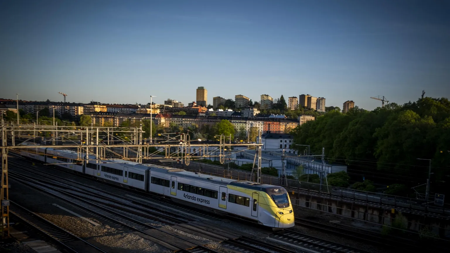 Ett tåg på ett järnvägsspår med en stadssilhuett och träd i bakgrunden under en klar himmel.