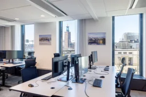 Ett kontor med datorer på vita skrivbord, stora fönster och utsikt över en stad med ett torn i bakgrunden.