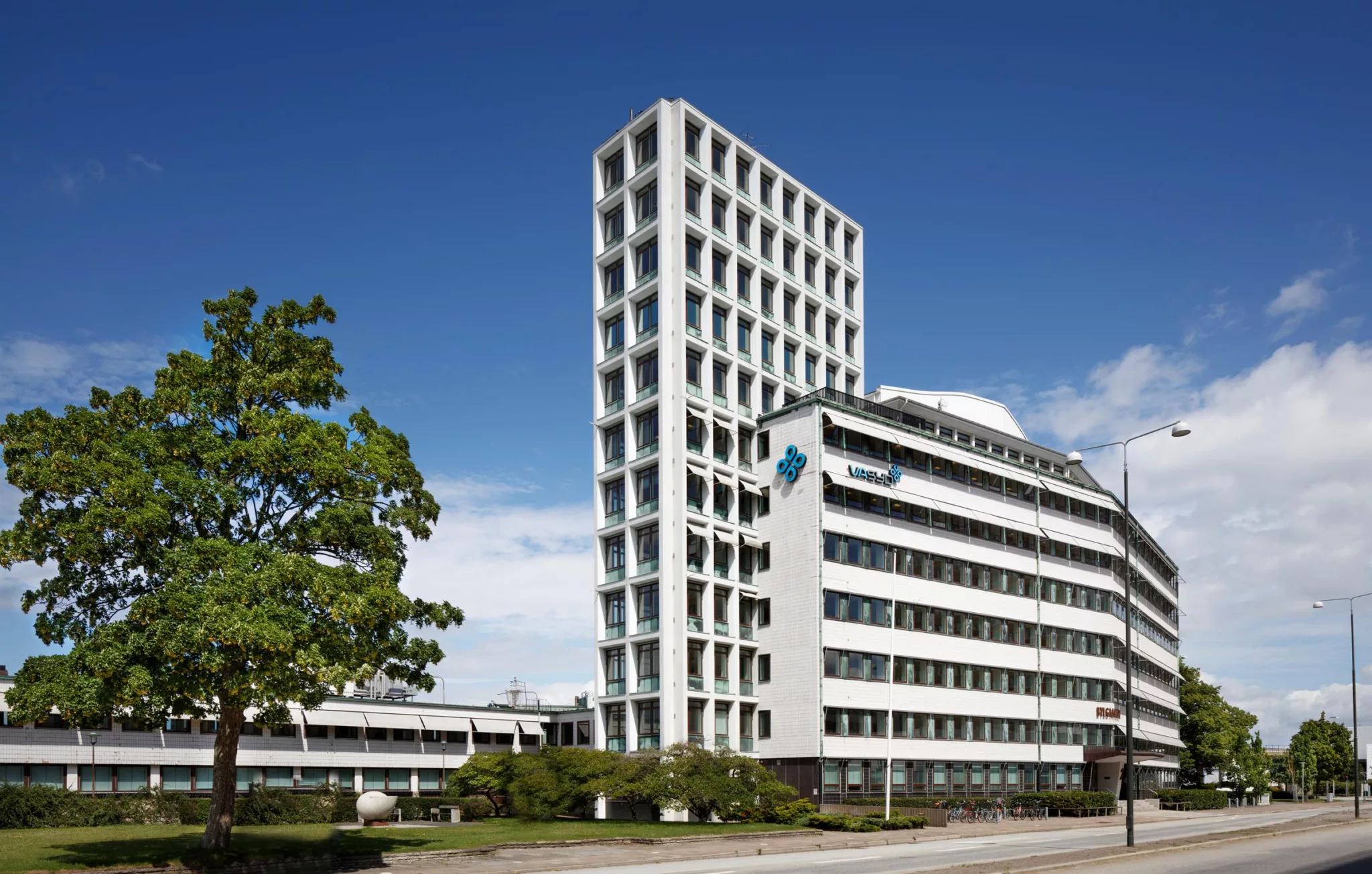 Höghus med modern fasad och flera fönster bredvid en lägre byggnad. Träd och grönska i förgrunden under blå himmel.