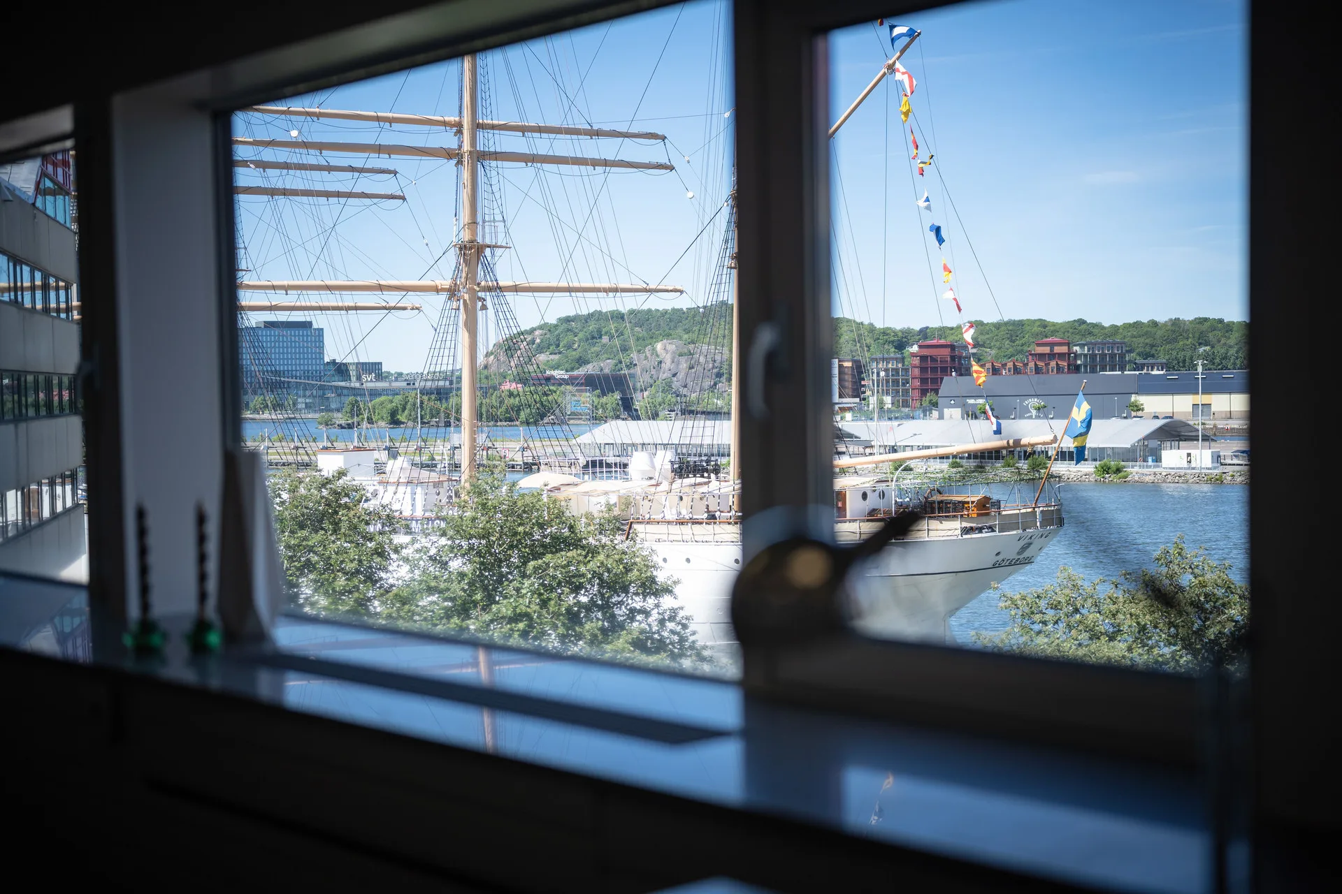 Utsikt genom ett fönster mot en hamn med en segelbåt och byggnader, med detaljer från fönsterkarmen i förgrunden.