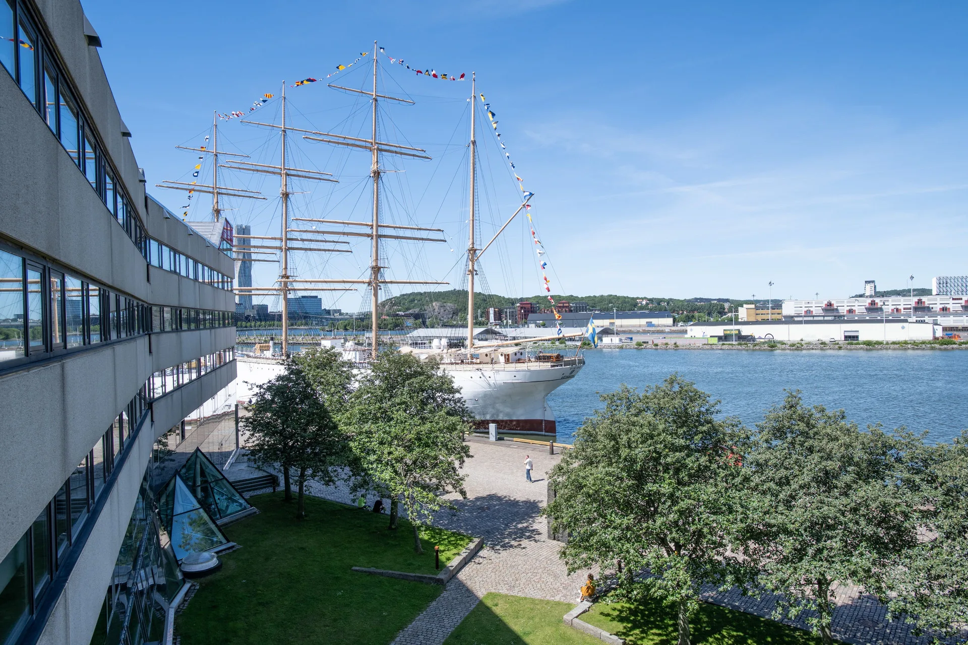 En stor segelbåt vid en hamn, omgiven av grönska och byggnader under en klarblå himmel.