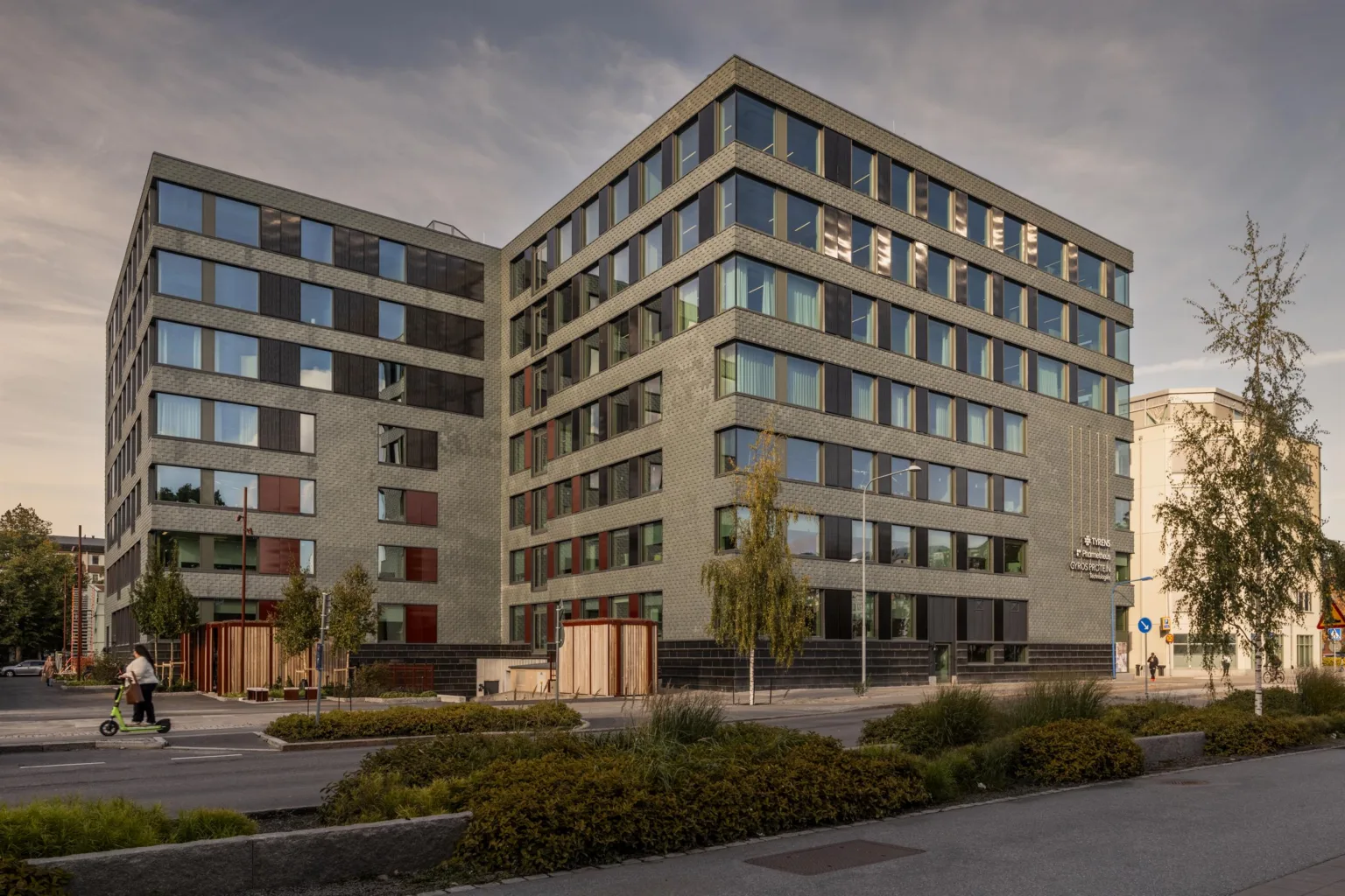 Exteriör av ett flerfamiljshus med flera våningar och stora fönster, omgiven av grönområden.
