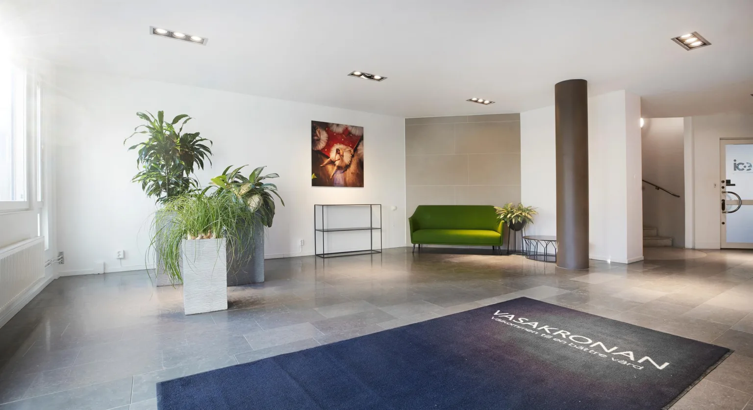 En modern reception med en grön soffa, växter i krukor, en konsttavla på väggen och en mörk matta med text.