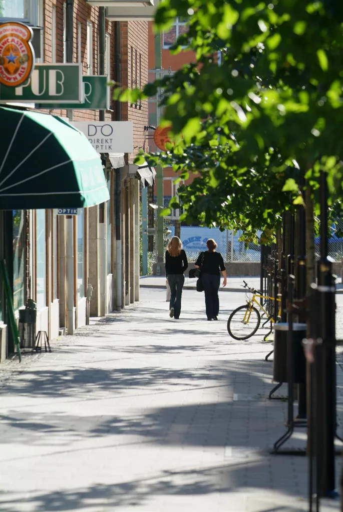 Två personer går längs en solig trottoar kantad av träd och butiker.