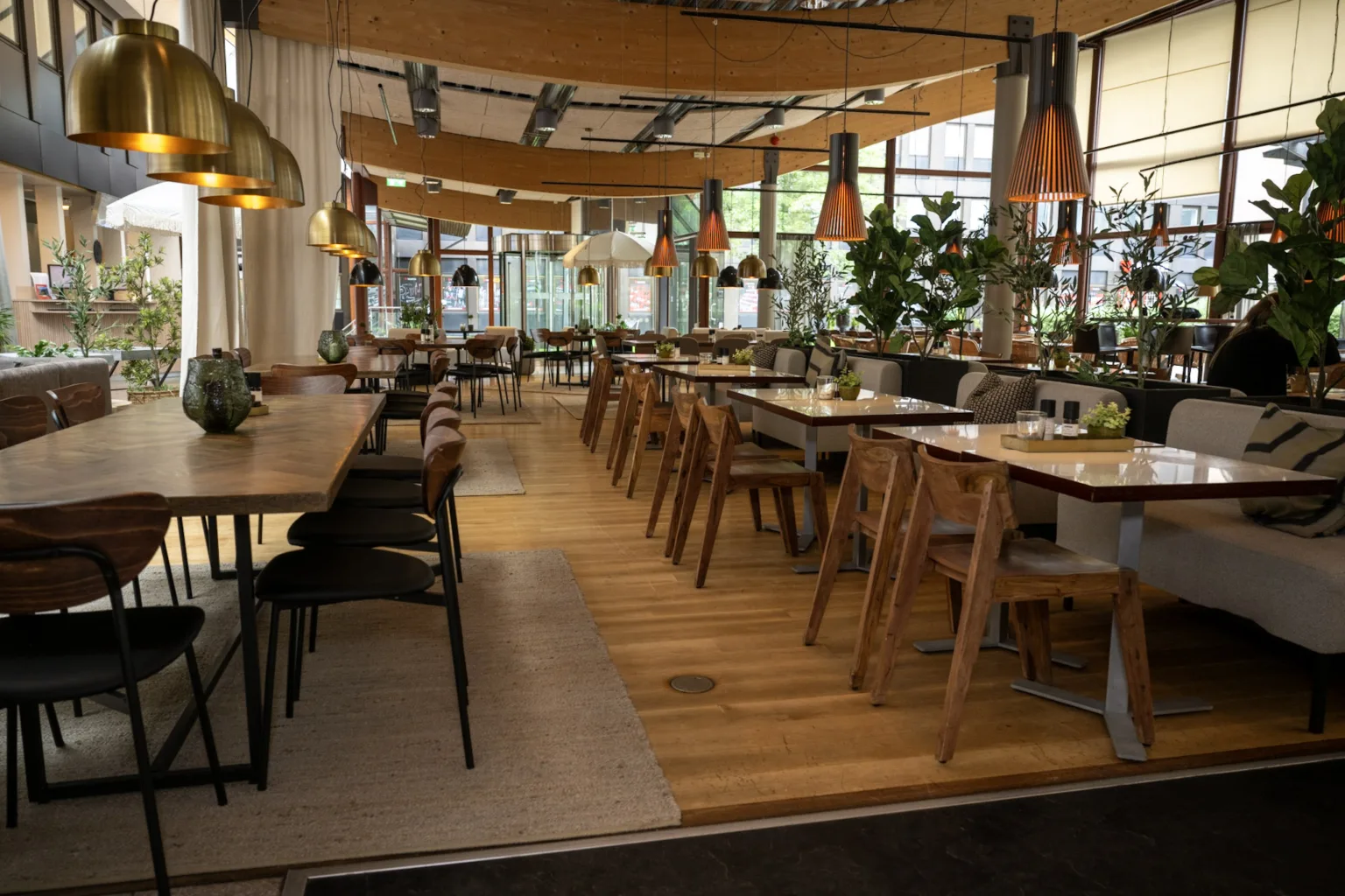 En restaurang med träbord, stolar och stora fönster som släpper in naturligt ljus; en inbjudande och grön interiör.