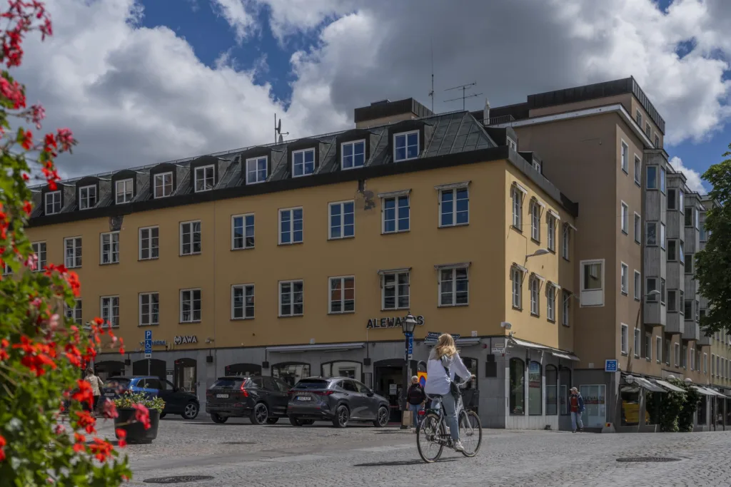 En gul byggnad med flera fönster och personer som cyklar och promenerar på en solig dag, med moln på himlen.