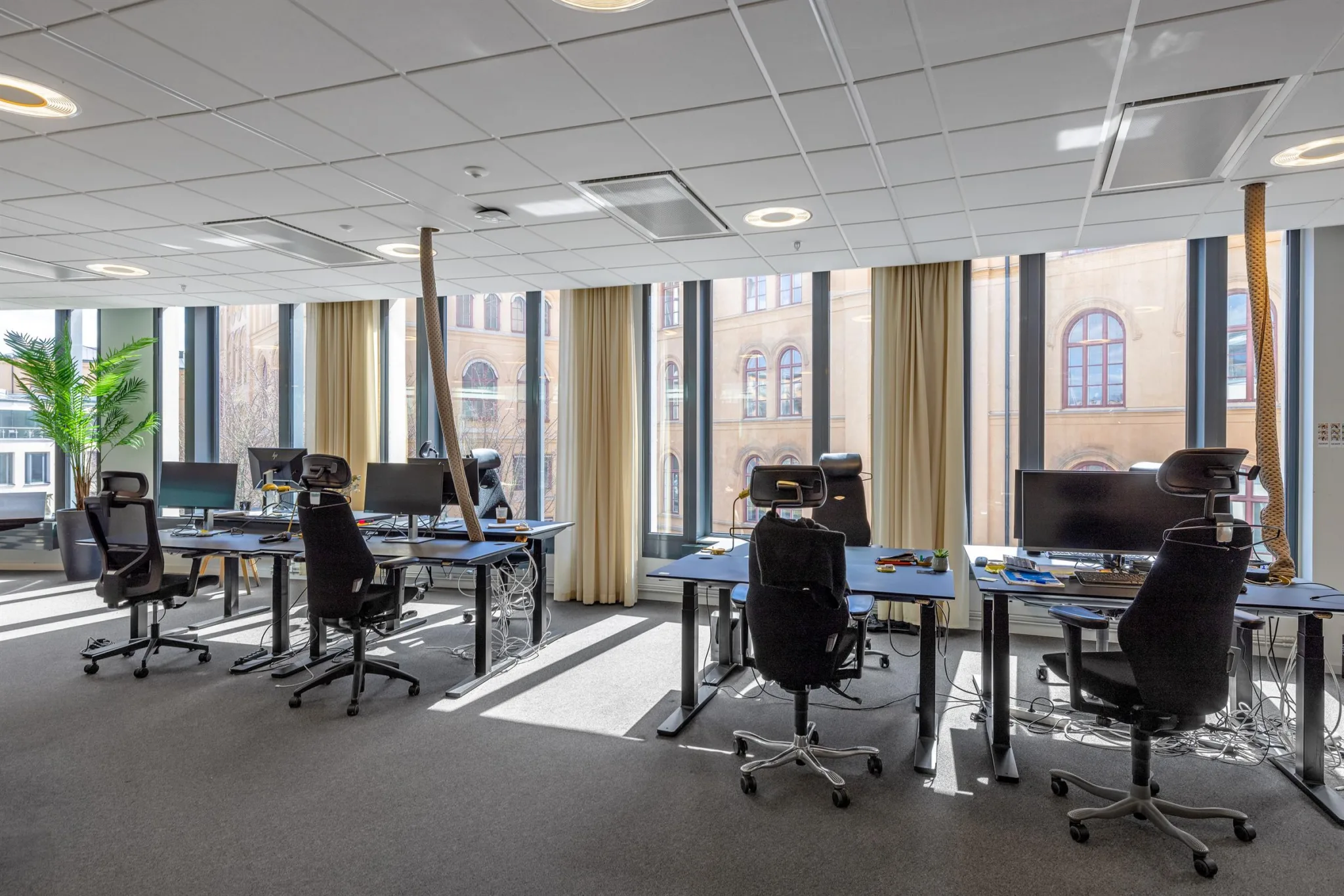 3. Ett kontorsrum med flera skrivbord och datorer, stora fönster med gardiner.