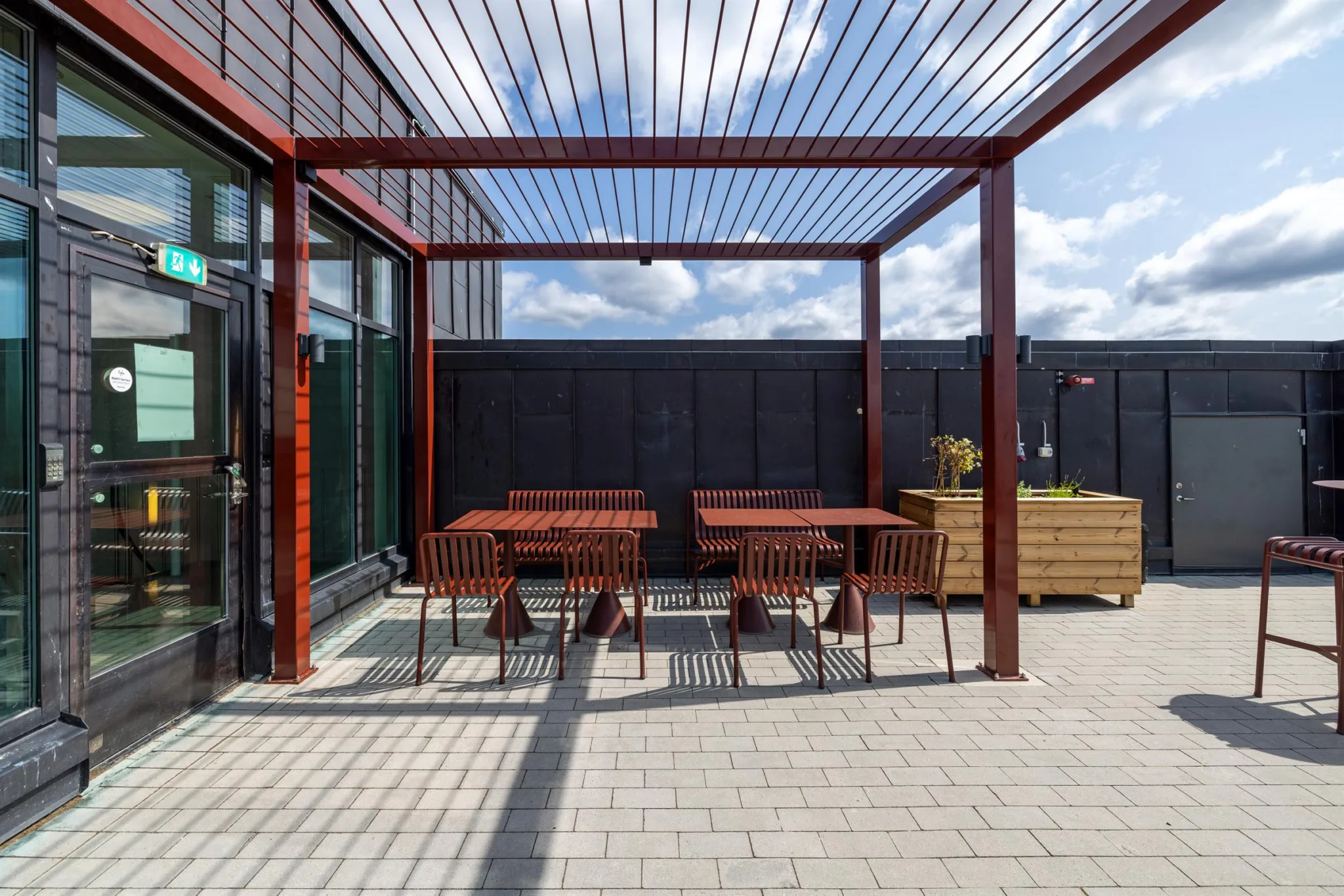 1. En utomhusterrass med två röda bord och stolar under en pergola, med en plantering i bakgrunden och en blå himmel ovanför.