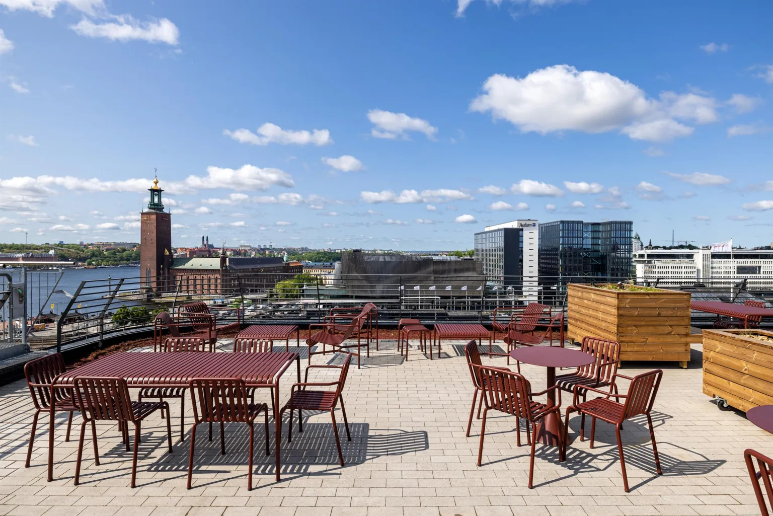 En takterrass med röda bord och stolar, med vy över Stadshuset och moderna byggnader.