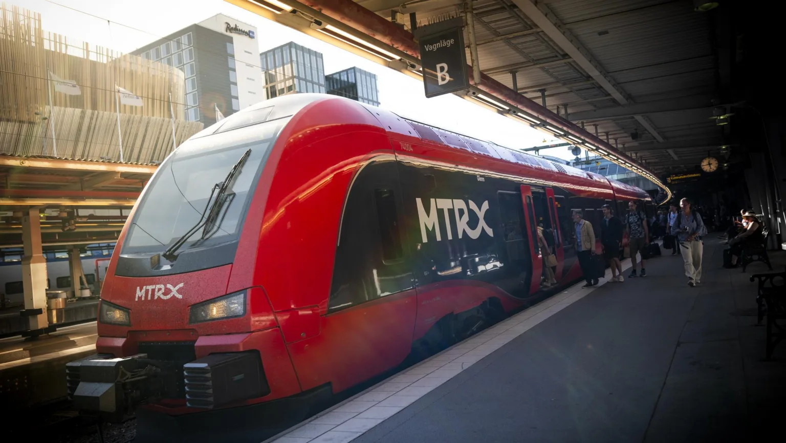 Ett rött MTRX-tåg står vid en perrong på en tågstation.