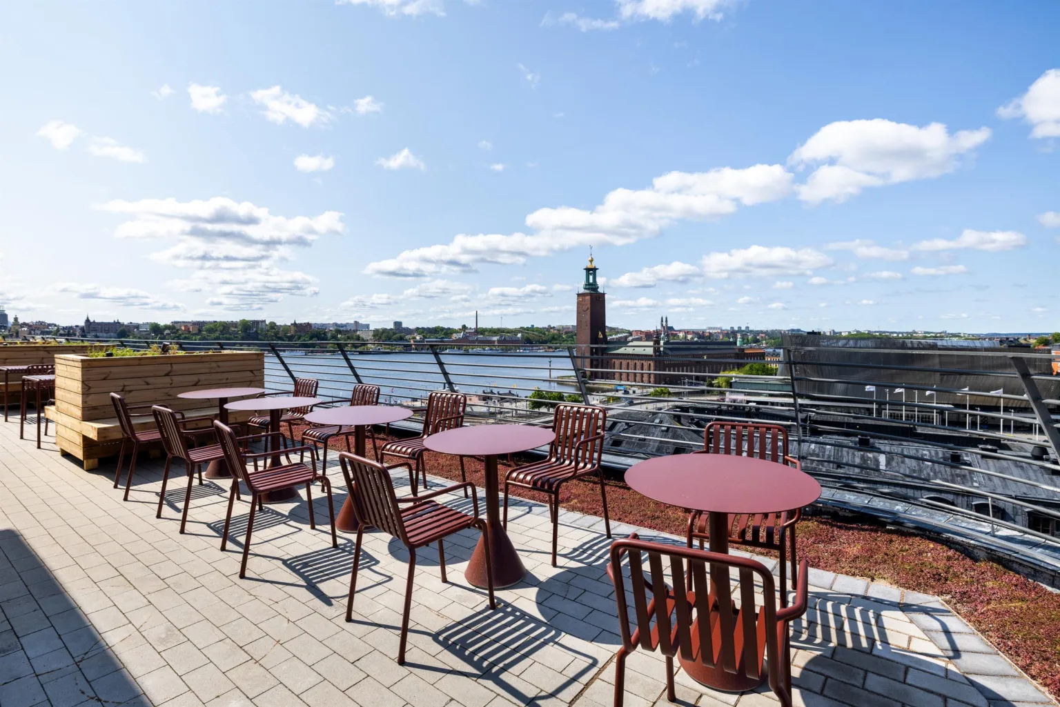 En takterrass med röda bord och stolar, med utsikt över en sjö och Stadshuset i bakgrunden.