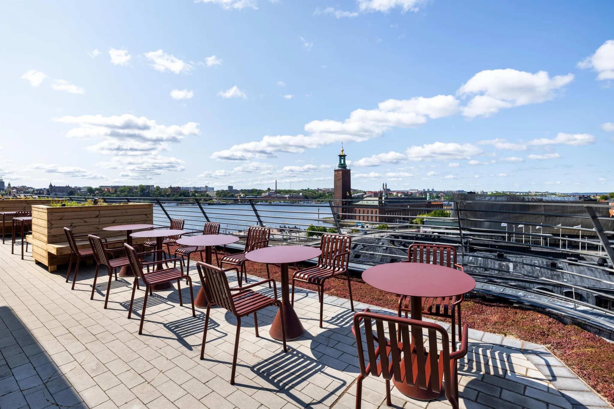 En takterrass med röda bord och stolar, med utsikt över en sjö och Stadshuset i bakgrunden.
