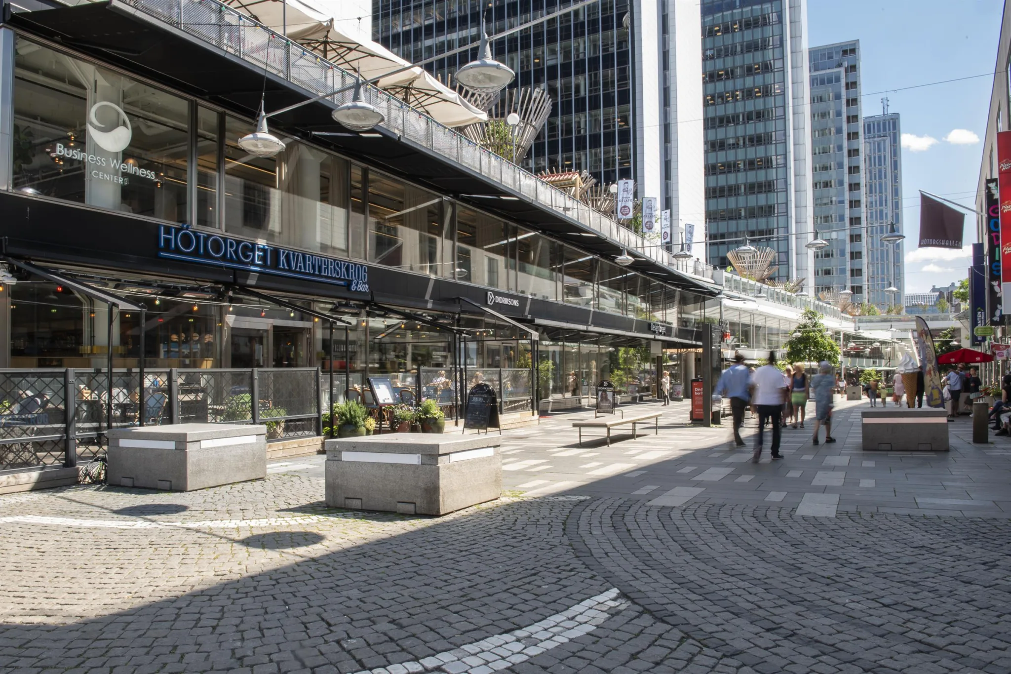5. En bred gångväg omgiven av höga byggnader, med butiker, planteringar och människor som promenerar.