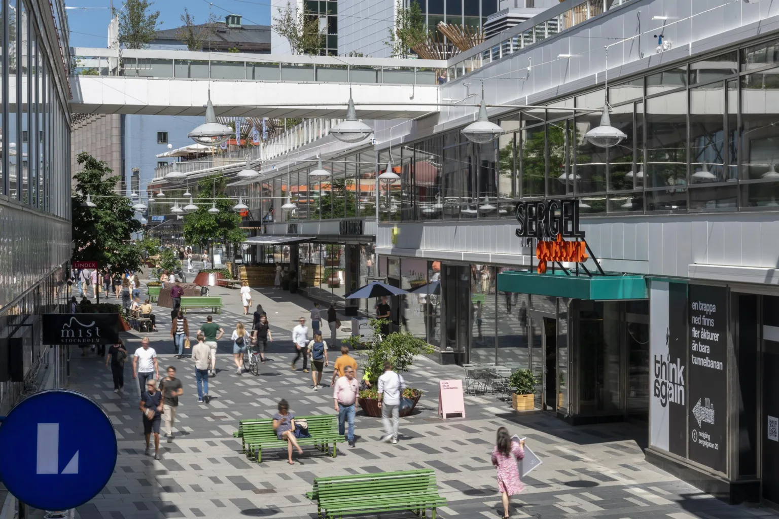 1. En gågata med människor som promenerar och butiker på båda sidor, omgiven av moderna byggnader och grönska.