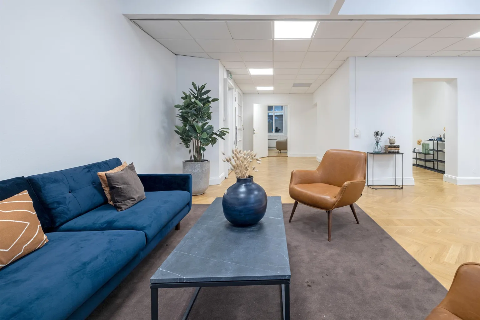 2. Ett vardagsrum med en blå soffa, en brun fåtölj, ett grått soffbord med en blå vas och en stor grön växt.