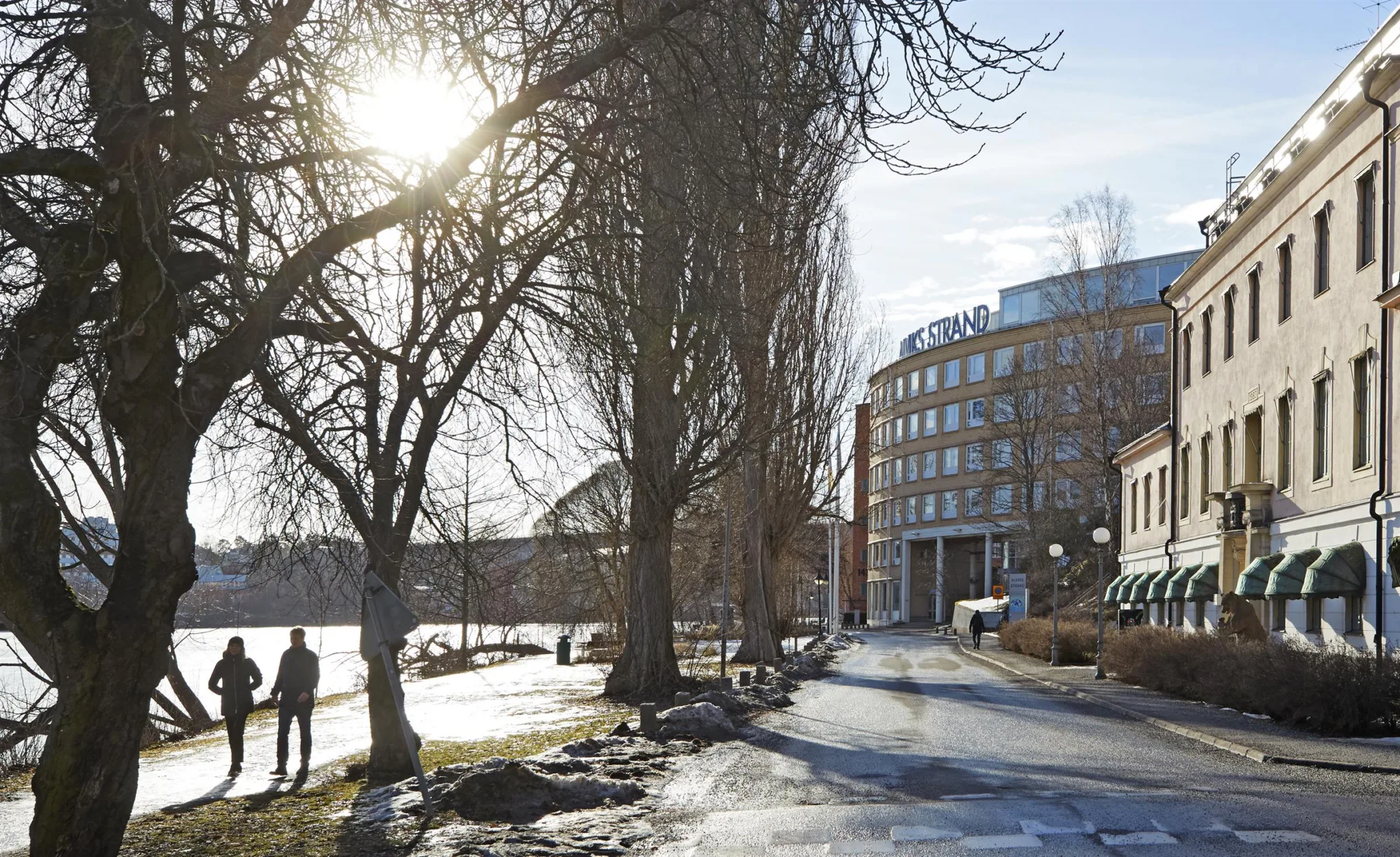 En gångväg med snöfläckar och träd längs sidorna, där solen skiner genom grenarna, med en byggnad i bakgrunden.