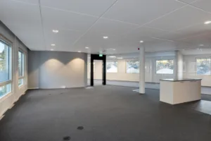 Ett tomt kontorsutrymme med grå heltäckningsmatta, vita väggar, stora fönster och infällda taklampor.