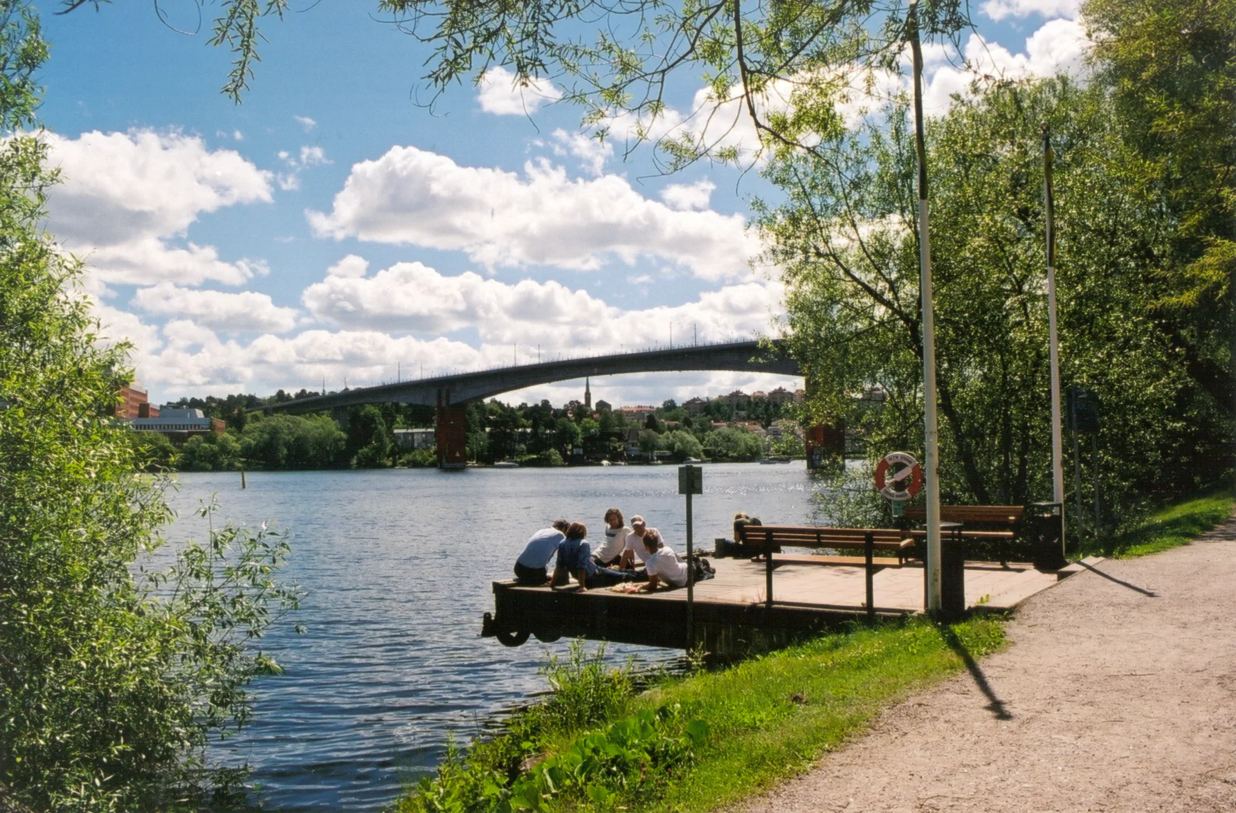 En brygga vid en sjö med människor som sitter och njuter av solen, omgiven av gröna träd och en bro i bakgrunden under en molnig himmel.