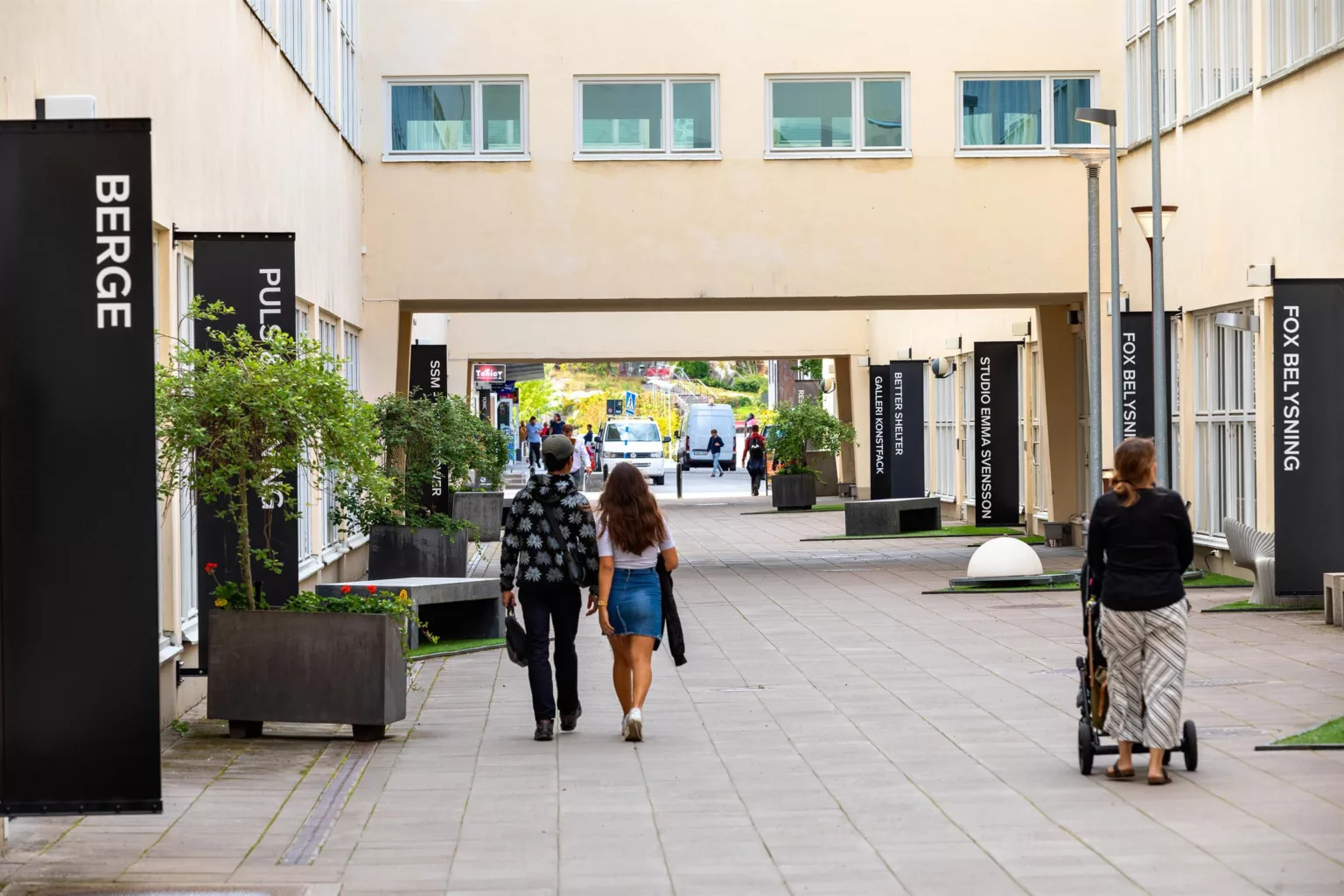 3. En gångväg mellan byggnader där människor promenerar, med gröna växter och skyltar längs sidorna.