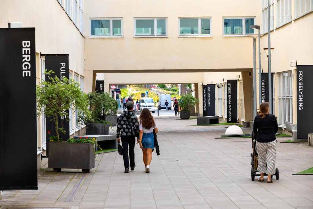 Gångväg mellan byggnader med människor som går, omgivna av växter och skyltar.
