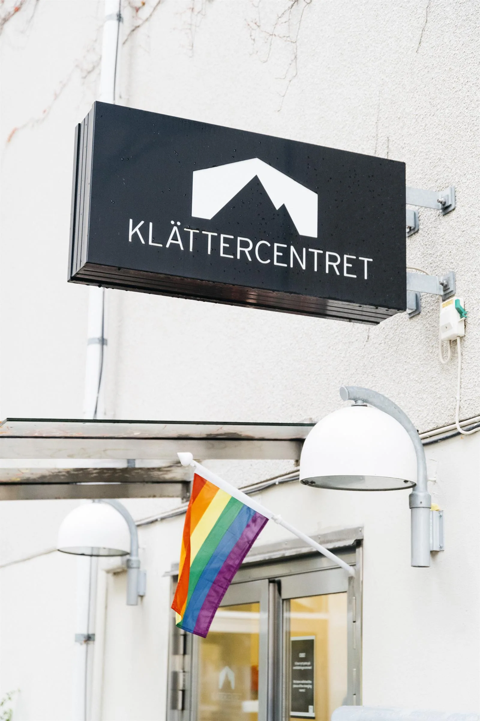 En svart skylt med texten "Klättercentret" och en vit bergsymbol ovanför, samt en regnbågsflagga som hänger under skylten.