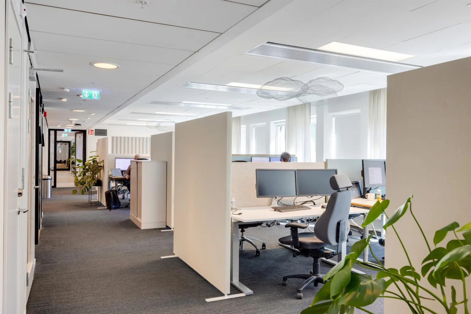 Ett ljust kontor med skrivbord, kontorsstolar, datorskärmar och gröna växter.