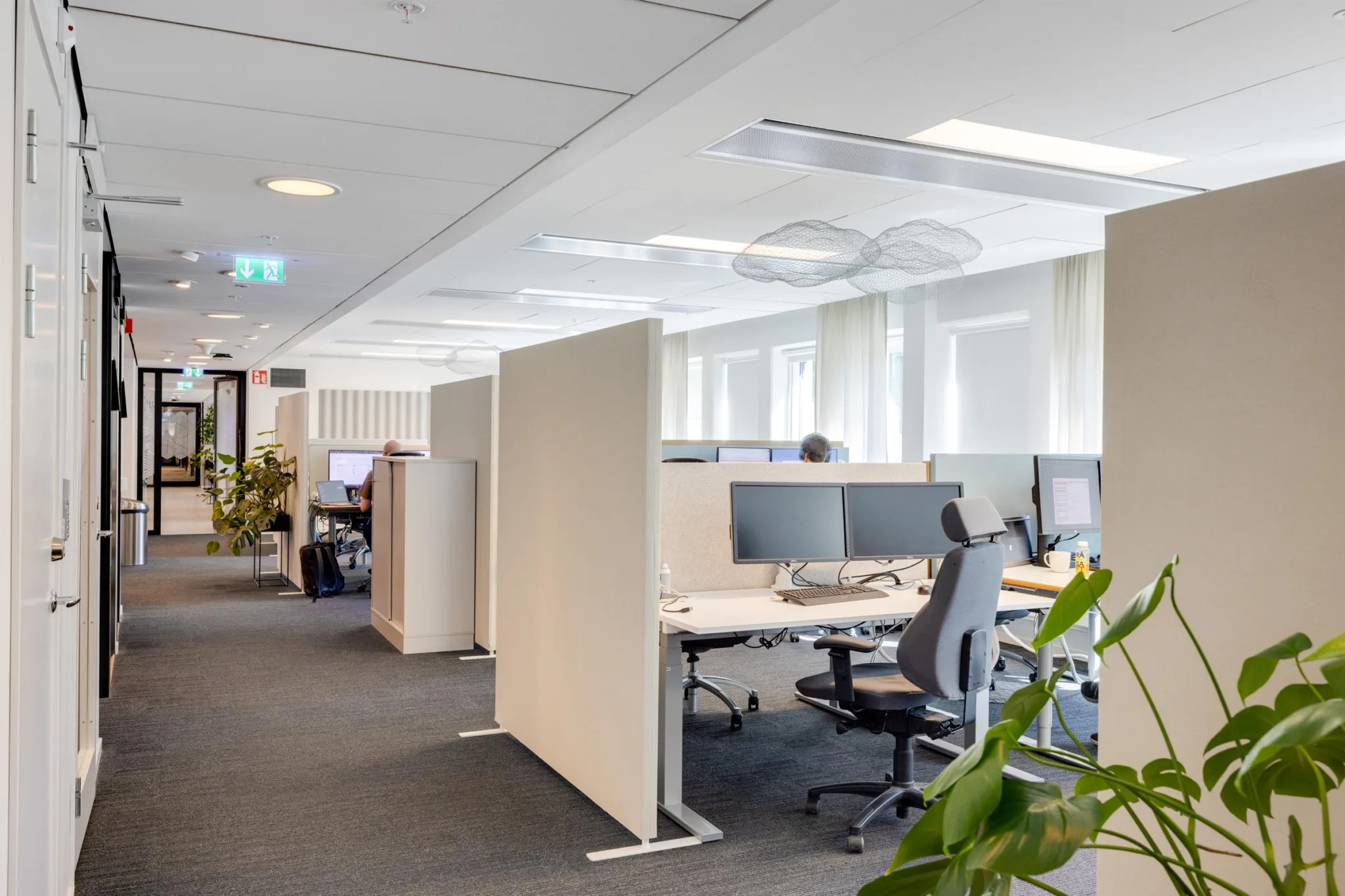 Ett ljust kontor med skrivbord, kontorsstolar, datorskärmar och gröna växter.