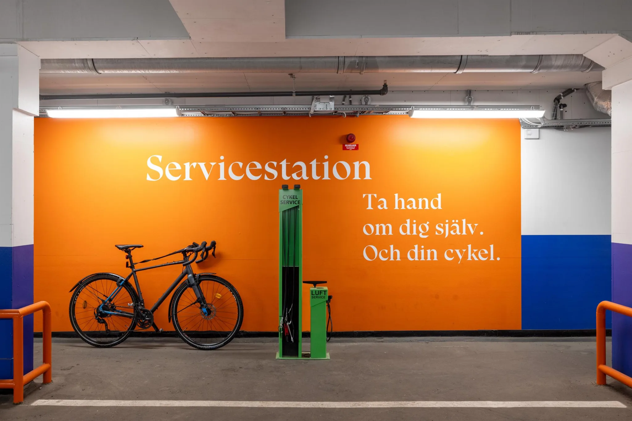 En orange vägg med texten "Servicestation. Ta hand om dig själv. Och din cykel." bredvid en cykel och en grön cykelpump.