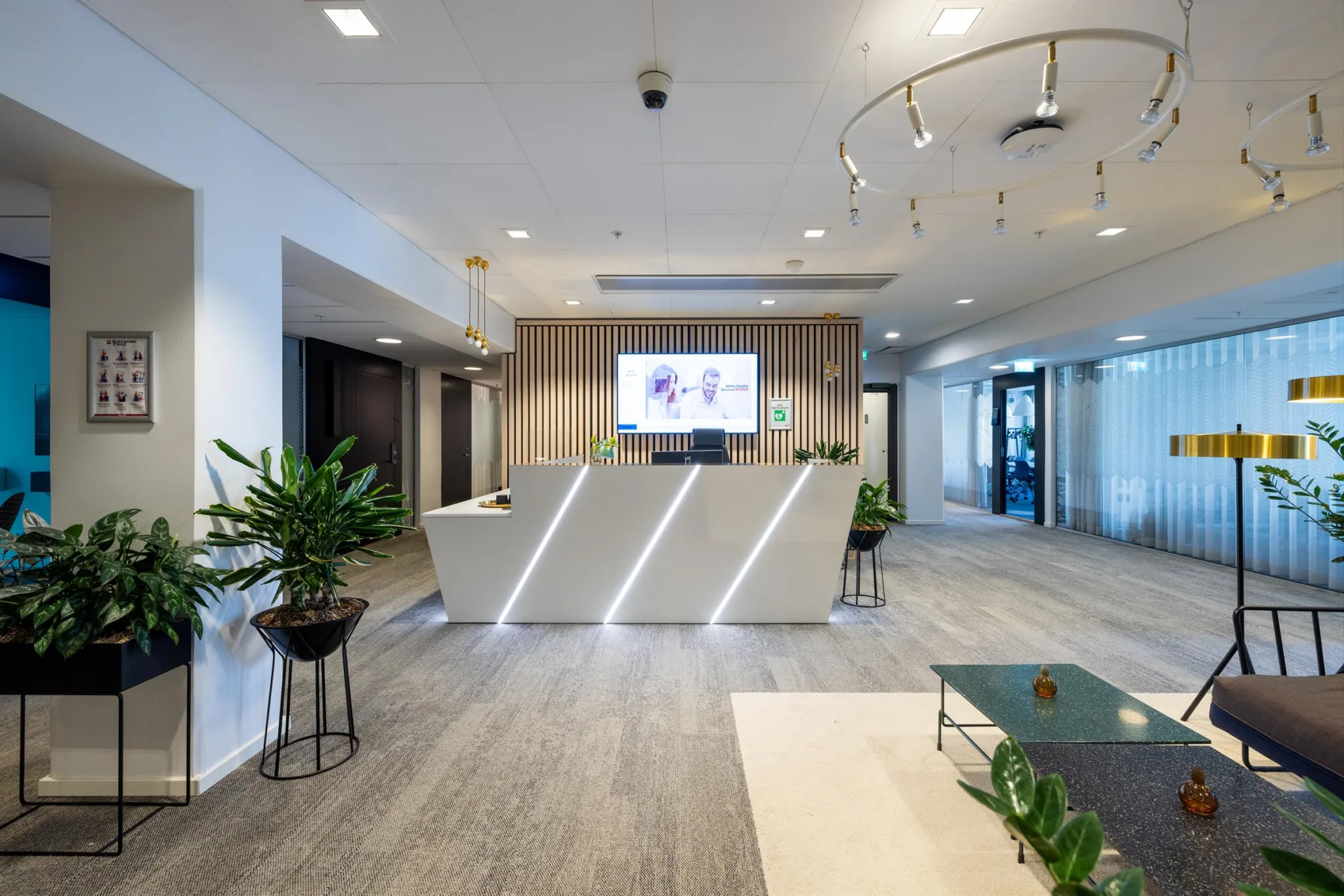 En reception med en vinklad vit disk, gröna växter och en stor skärm på väggen.