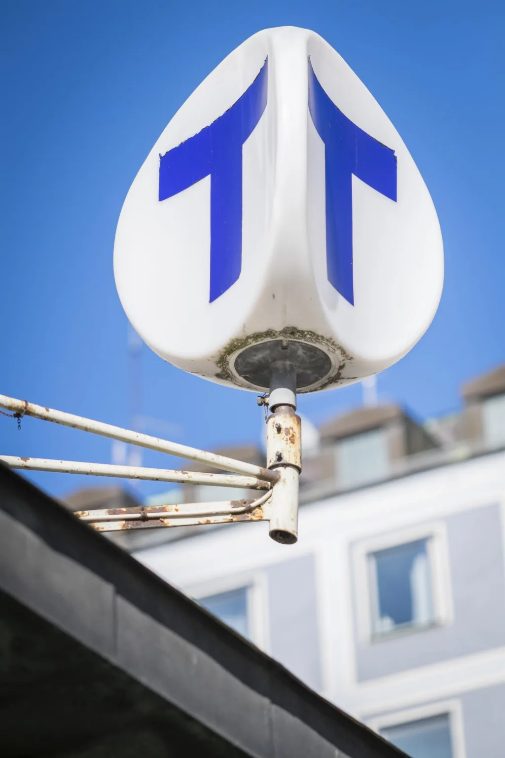 En vit vägskylt med två blå pilar som pekar uppåt, monterad på en stolpe mot en blå himmel och byggnader i bakgrunden.