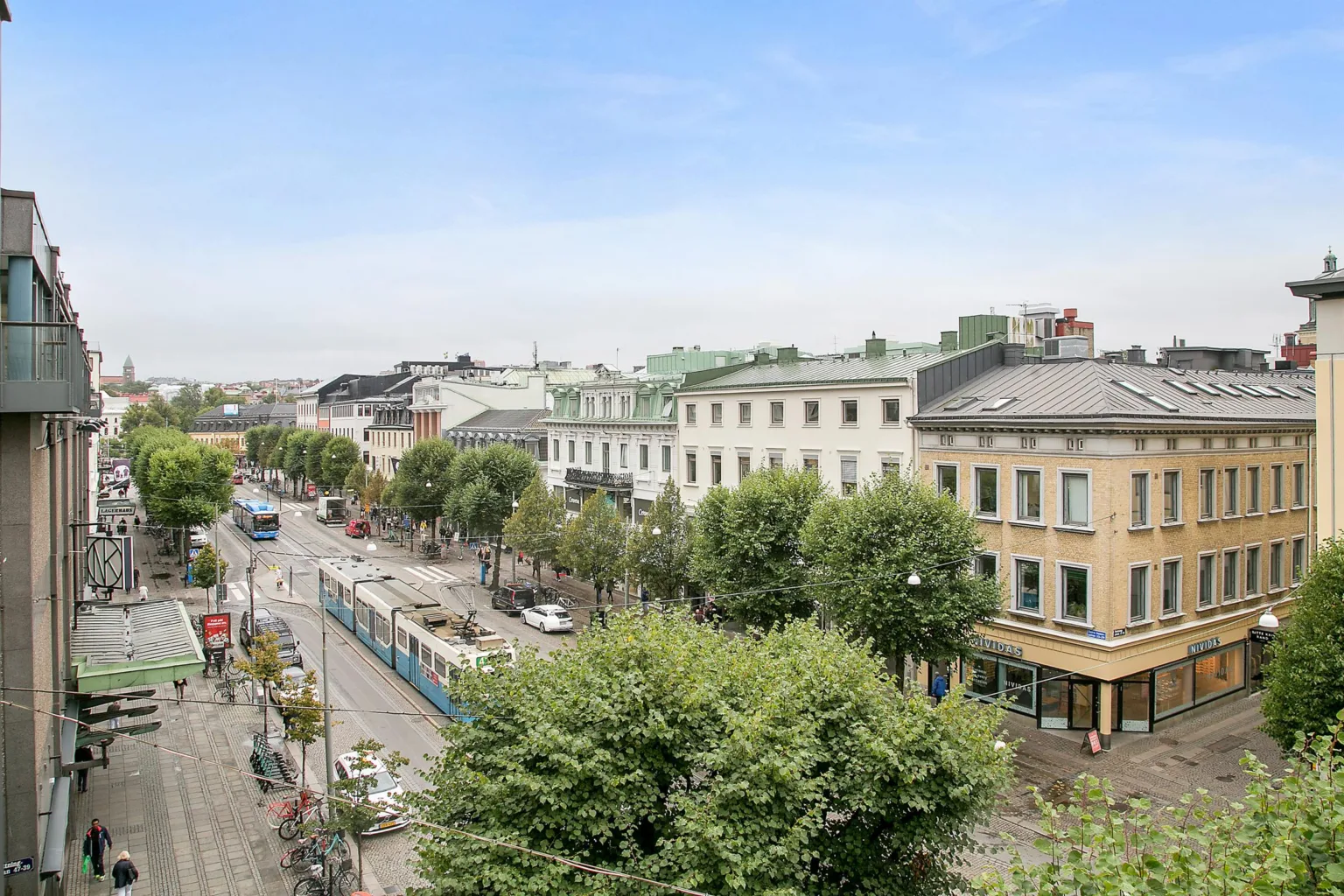 En stadsvy med en gata omgiven av träd och byggnader, samt en spårvagn och gående.