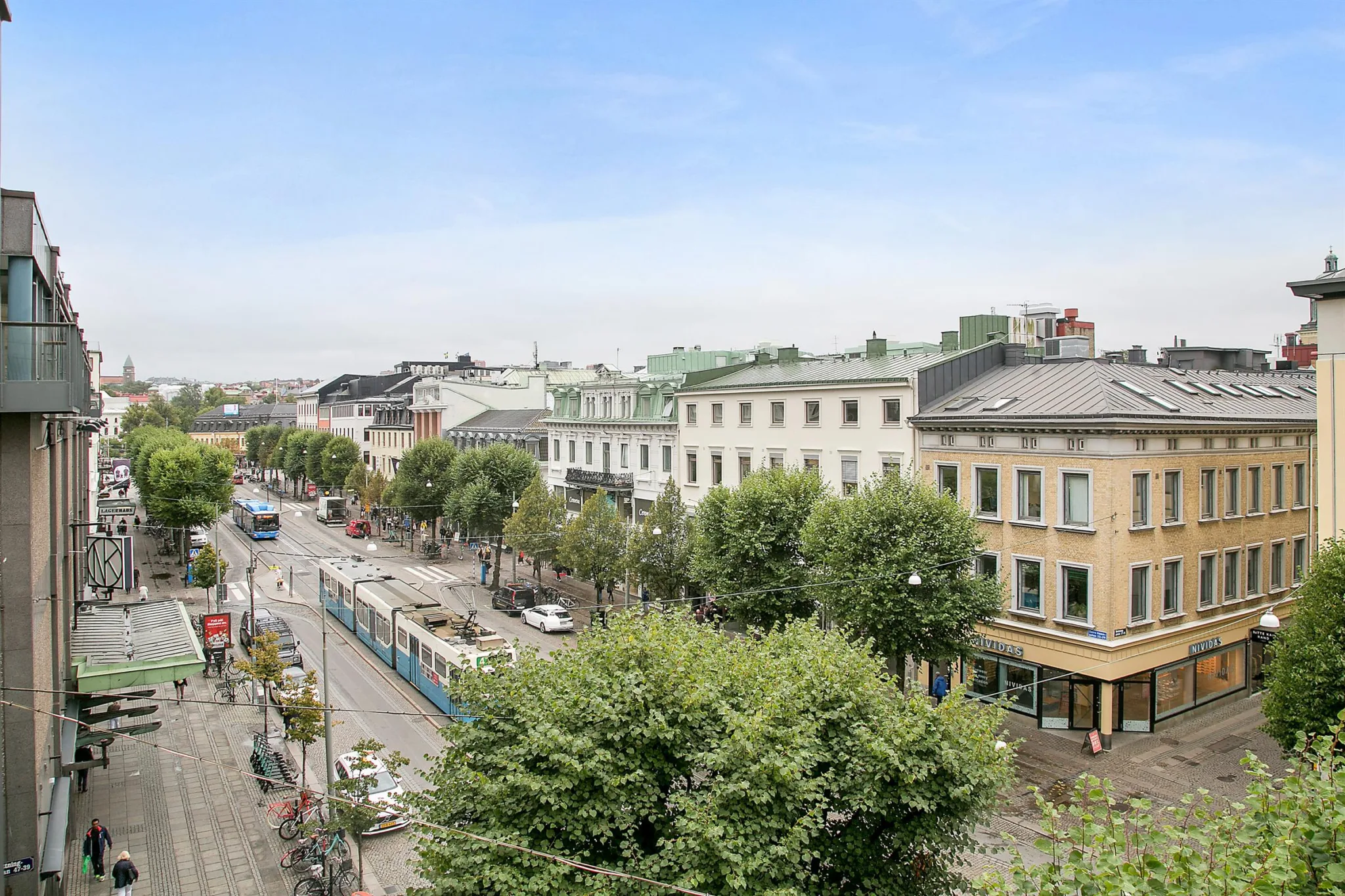 En stadsvy med en gata omgiven av träd och byggnader, samt en spårvagn och gående.