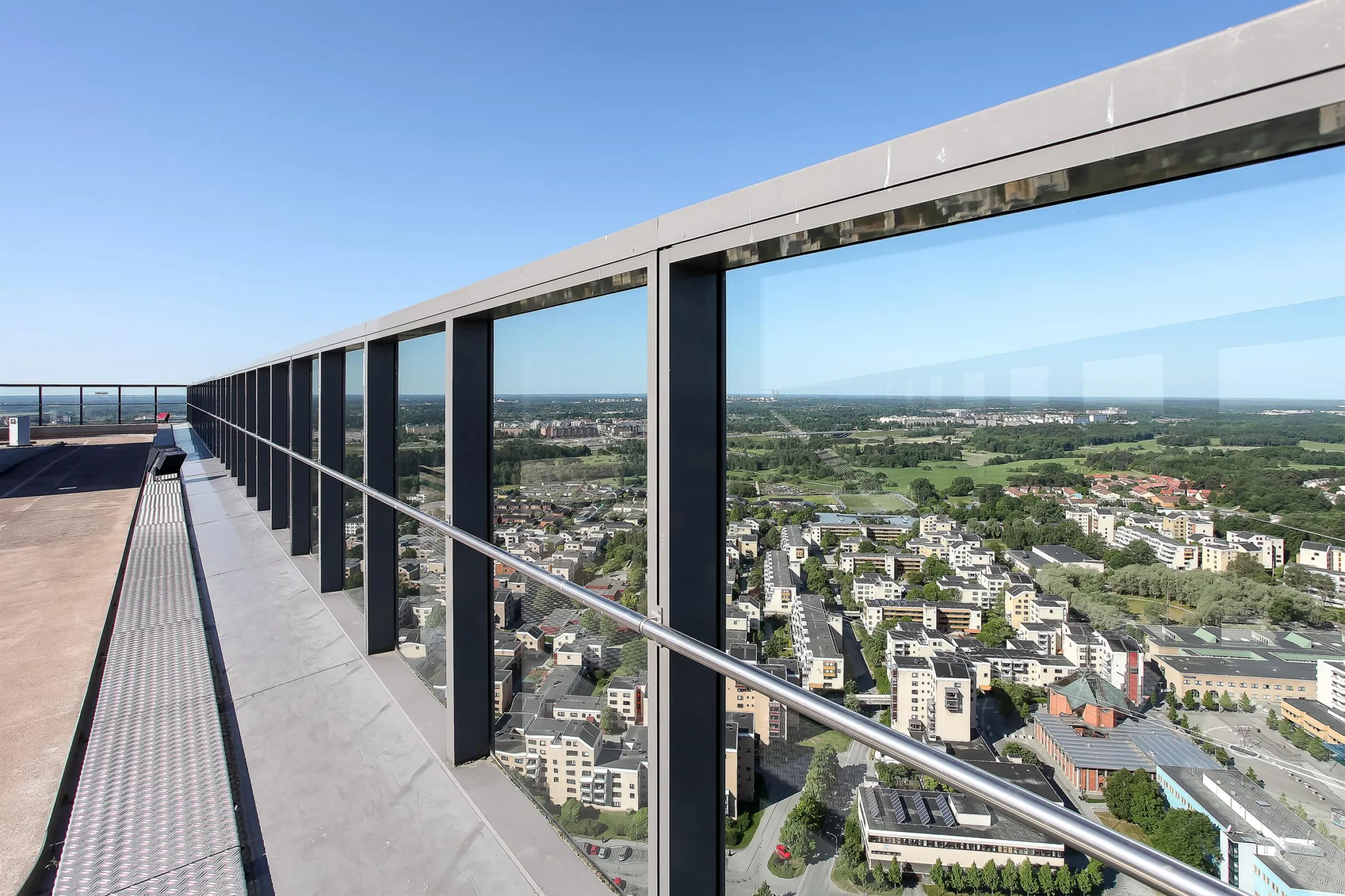 En balkong med glasräcke och utsikt över en stad med bostadsområden och grönska.