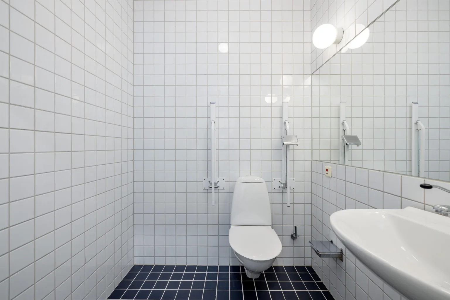 4. Ett rent och enkelt badrum med vita kakelväggar, svart golv, toalett, handfat och spegel.