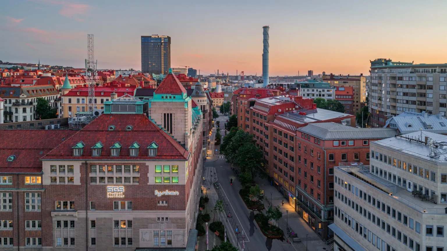 En stadsvy vid solnedgång med byggnader i rött tegel, en bred gata med träd, och en hög skorstensliknande struktur i bakgrunden.
