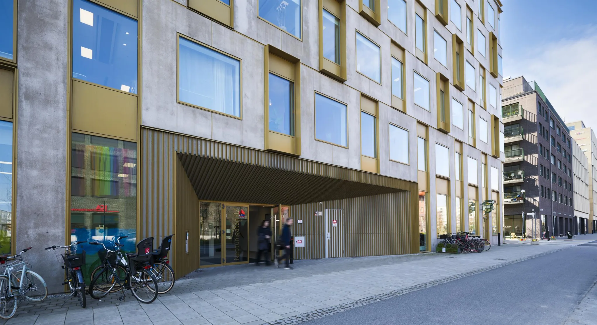 En modern byggnad med stora fönster och en ingång under en överhängande struktur, cyklar står parkerade utanför.