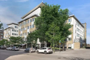En modern flerbostadsbyggnad med vita och beige fasader, omgiven av träd och parkerade bilar.