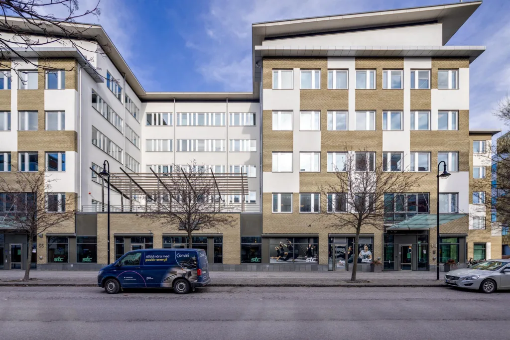 En byggnad med vit och gul fasad, sett från gatan med en blå bil parkerad framför.