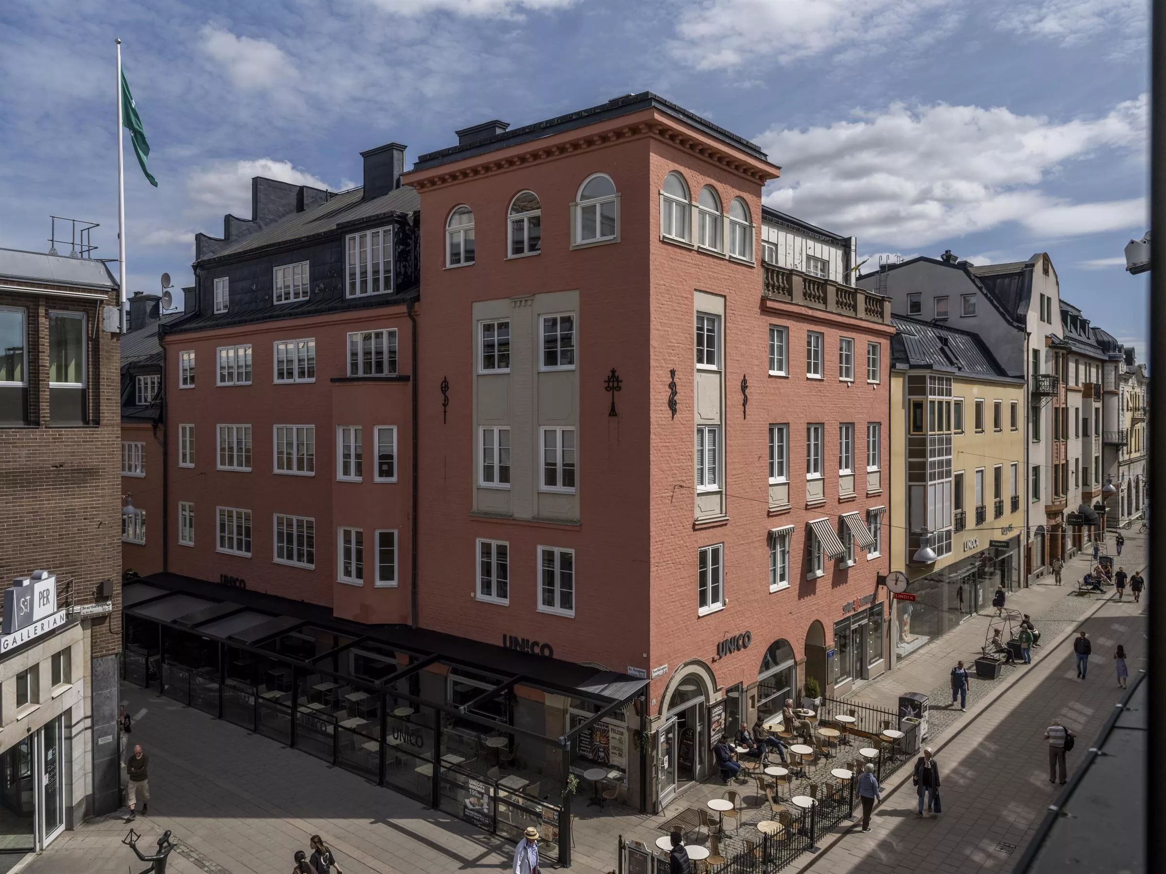 En rosa byggnad med flera våningar och uteservering vid trottoaren, omgiven av andra byggnader och fotgängare.