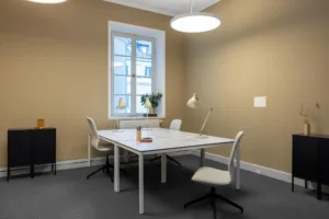 Ett litet kontorsrum med ett vitt bord, fyra stolar, en lampa och en blomvas vid fönstret.