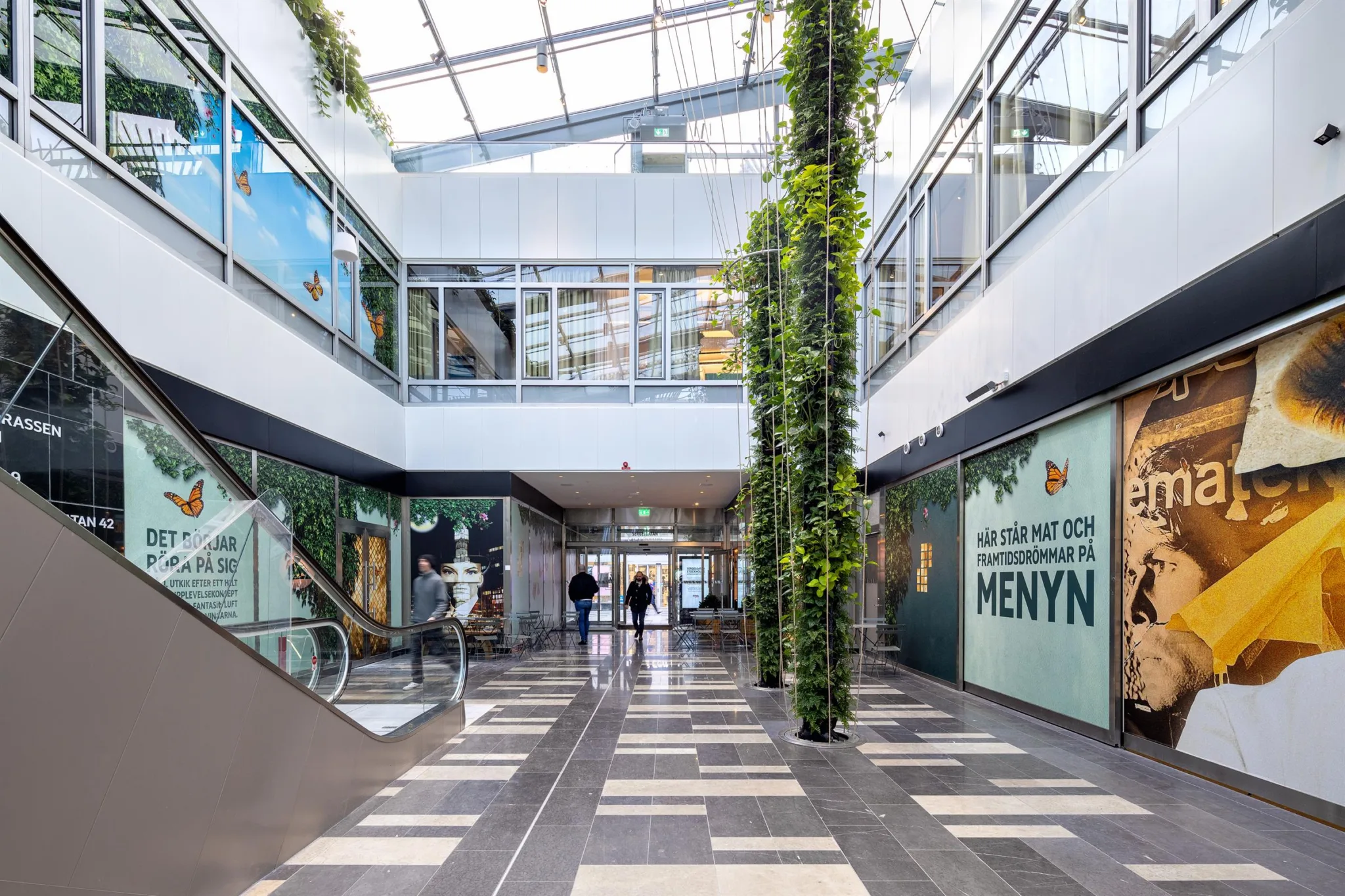 Inomhusgalleria med glasväggar, grönskande pelare, och skyltar för butiker och restauranger.