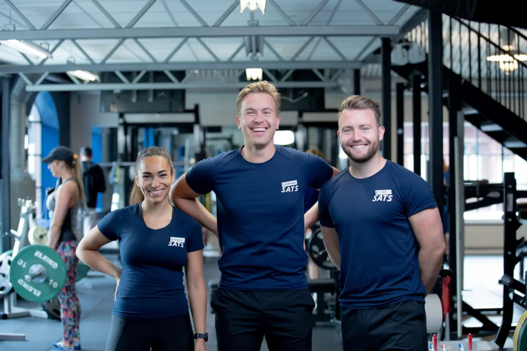 Tre personer i träningslokal, leende och klädda i mörkblå t-shirts med händer på höfter.