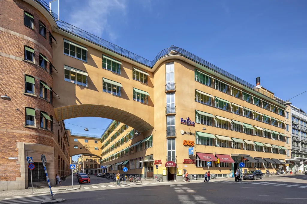 En gul byggnad med båge över gatan, flera skyltar och människor som går på trottoaren.