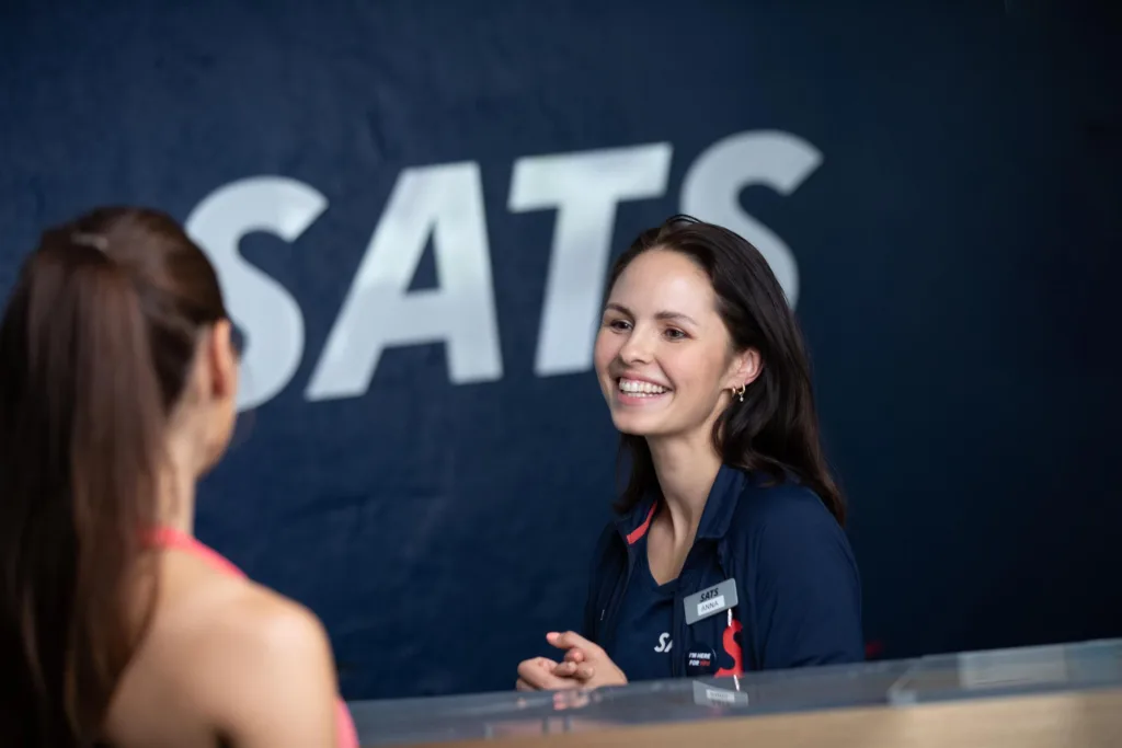En kvinna i en mörk uniform ler bakom en receptionsdisk med texten "SATS" på väggen.