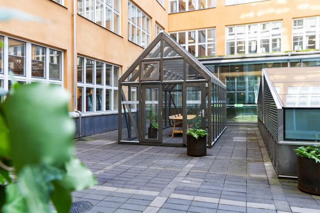 En innergård med ett växthus av glas och metall, omgiven av orange byggnader och planteringslådor.