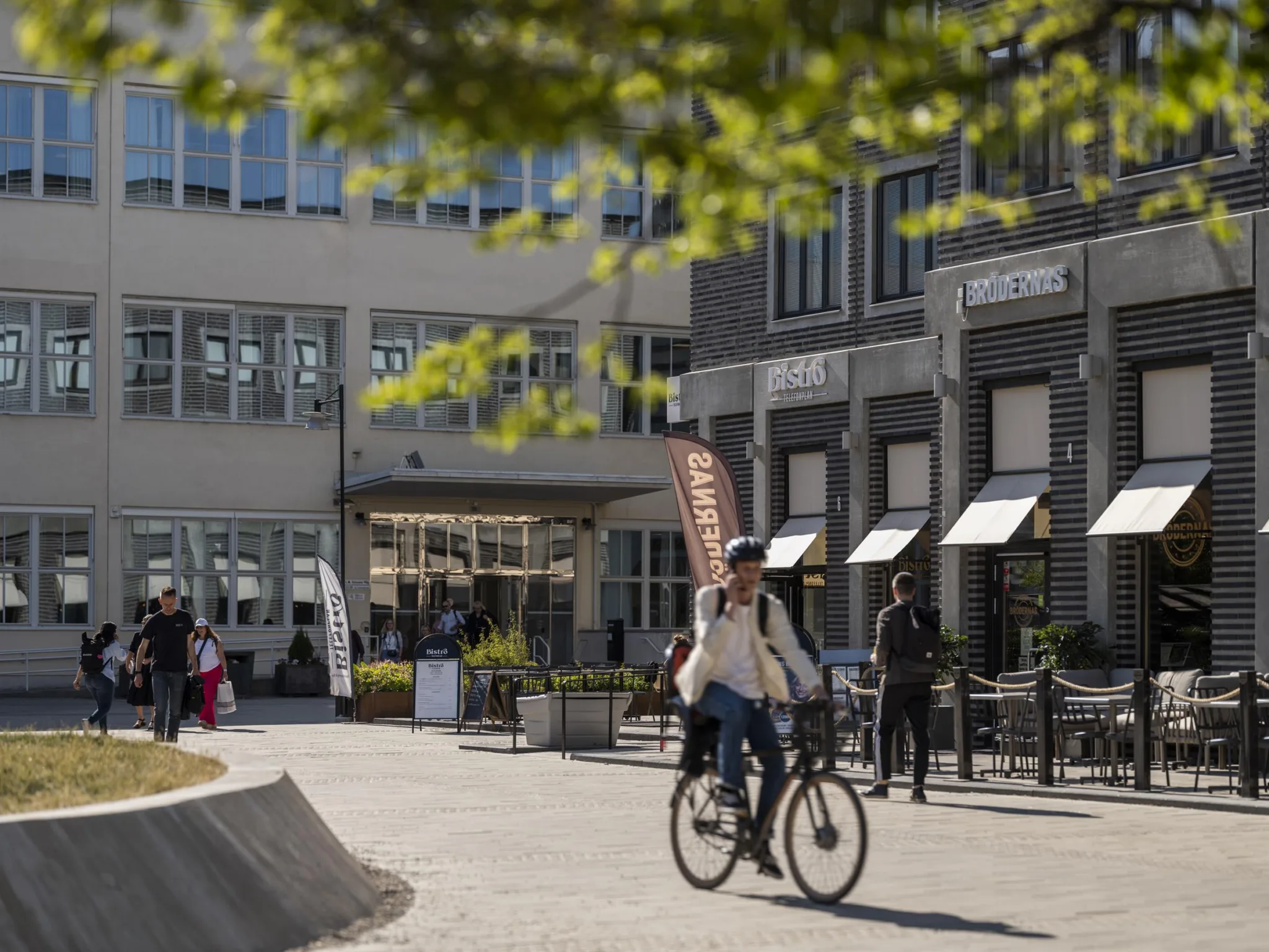 En person cyklar förbi en byggnad med flera butiker och människor som promenerar i solljus.