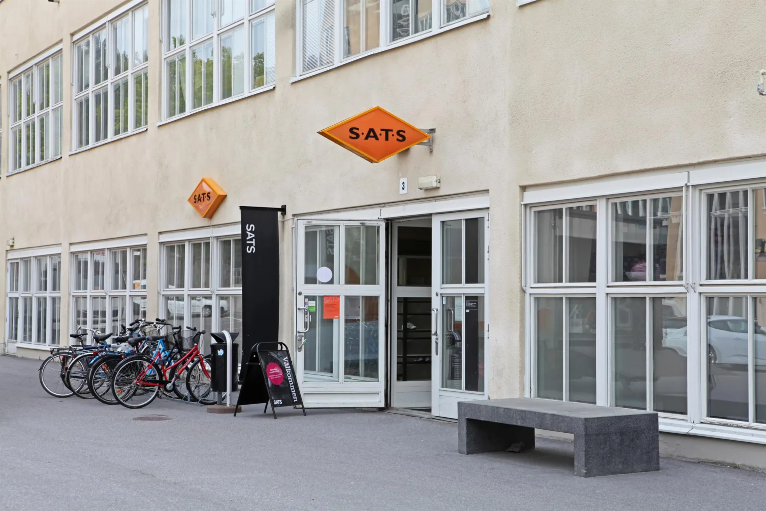 En byggnad med SATS-logga och entré, omgiven av cykelställ och bänkar.
